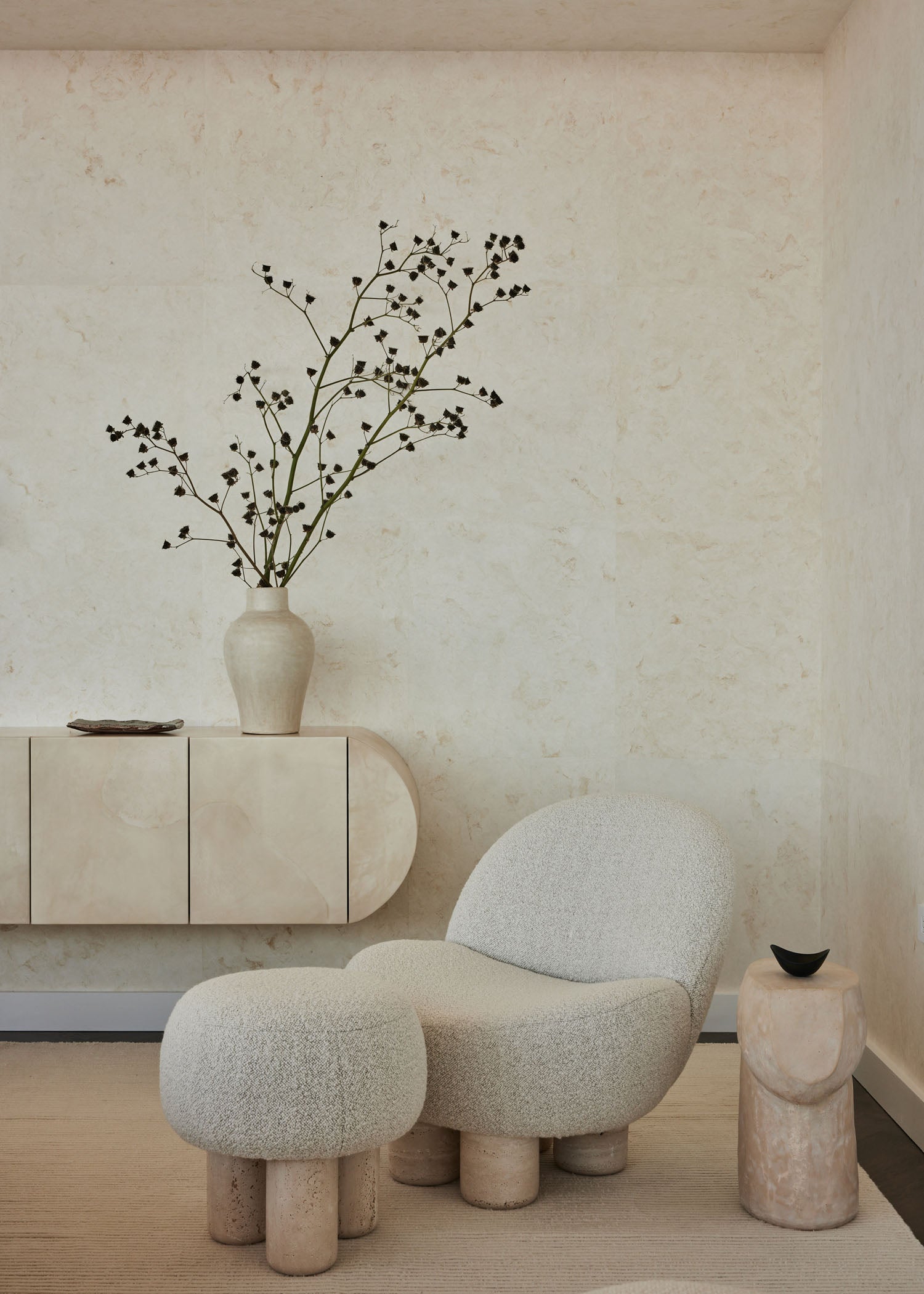 Beige armchair and ottoman with a vase of branches on a matching cabinet against a light wall.